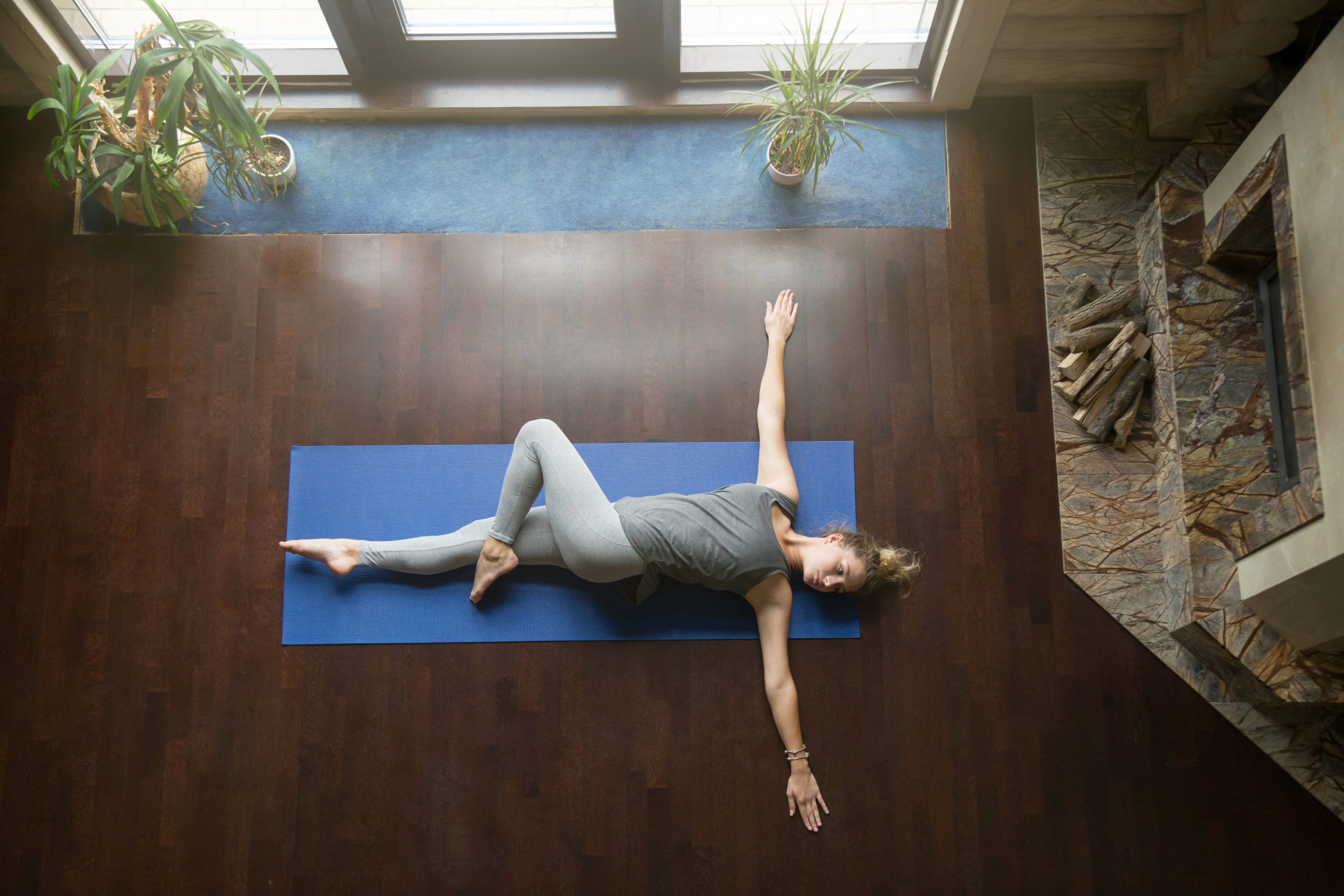 A woman performs a spinal twist stretch, while laying on a blue yoga mat in her home.