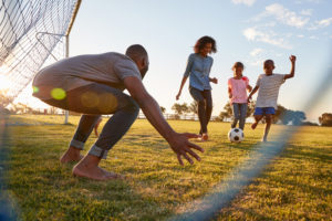 A mom and dad cheerfully play soccer with their children on a sunny afternoon. Dad playfully guards the goal while his son tries to score.