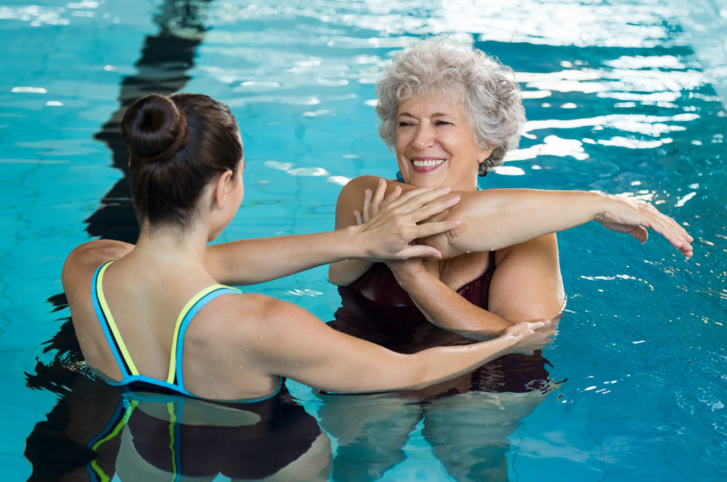 Young female therapist helping elderly woman stretch her arm in a pool during aquatic therapy.