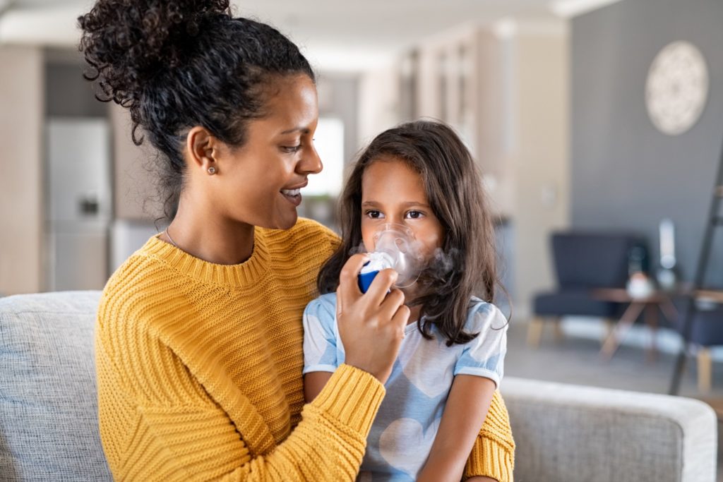 A mother in a yellow sweater is holding her daughter on her lap while administering an asthma breathing treatment using a nebulizer and child’s face mask.