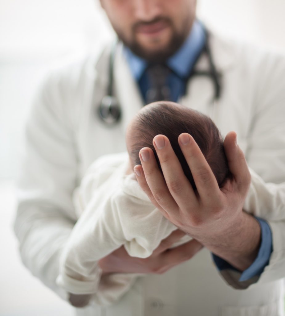 A close-up image of a doctor in a white jacket with a stethoscope around his neck cradling a newborn baby in his hands.