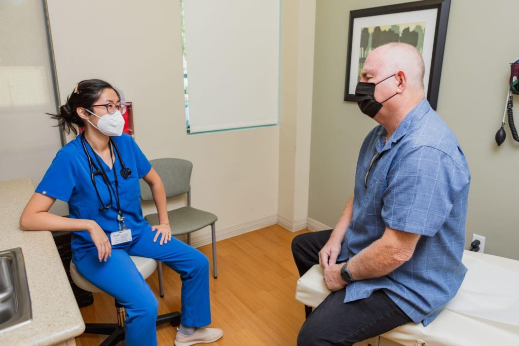 A male patient with a mask sits on an exam table across from a female doctor in blue scrubs in a mask, discussing care.