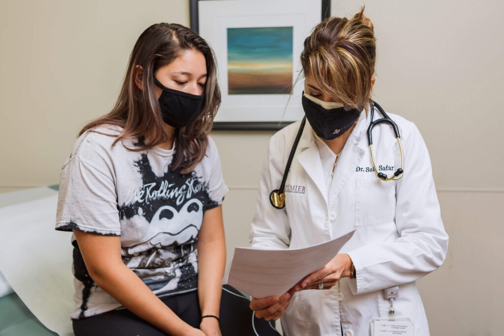 A young female patient with a mask sits on an exam table reviewing paperwork with a female doctor who is standing next to her.