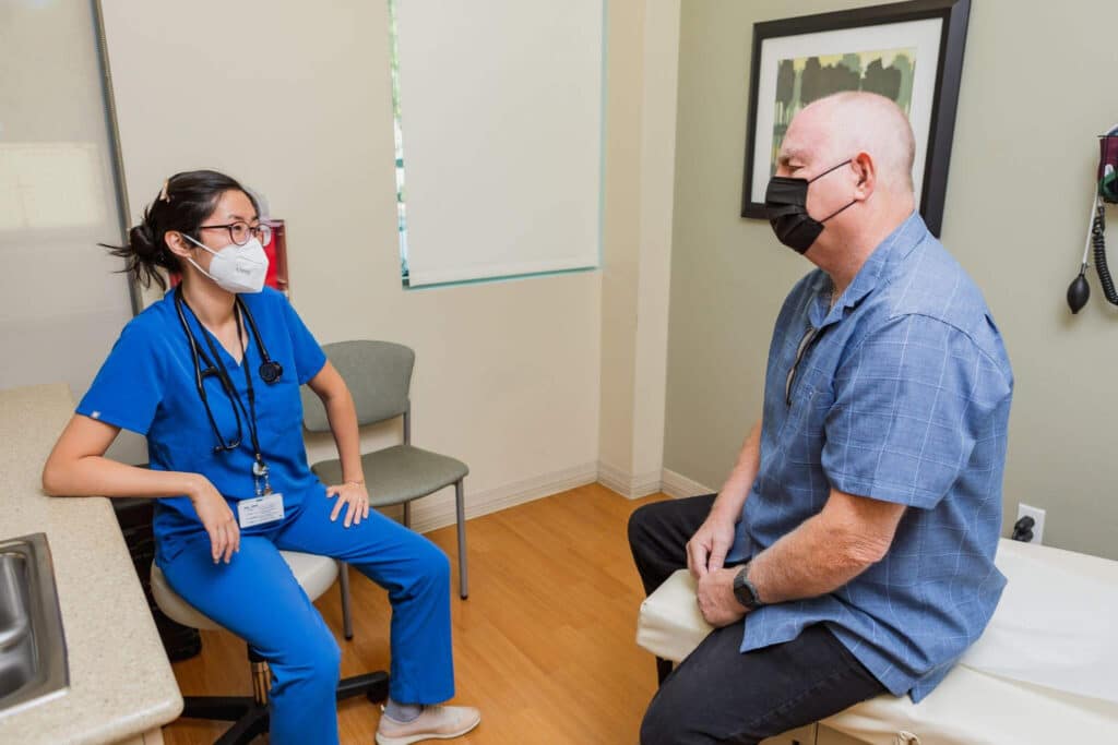 A female doctor in blue scrubs with a mask sits across from an older male patient on an exam table in a discussion.