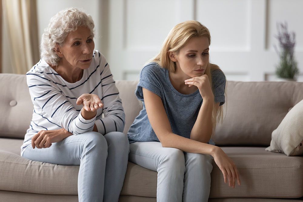 Unhappy woman in her forties sitting on the sofa next to her elderly mother with memory loss in her 70s.