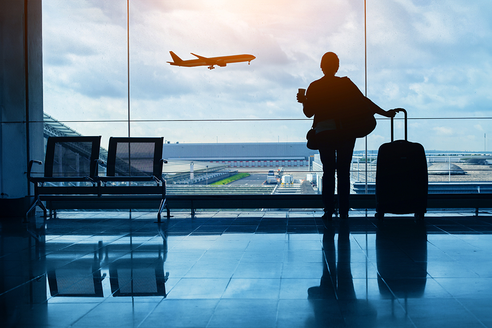 A woman with a suitcase stands in front of large windows watching a plane takeoff at an airport.