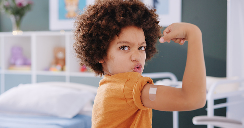 Young boy in a clinic proudly flexes his arm toward the camera, showing a small bandage where he received a vaccine.