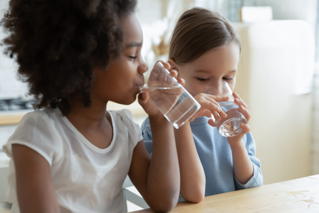 Two young girls drinking water out of glass cups over the sink.