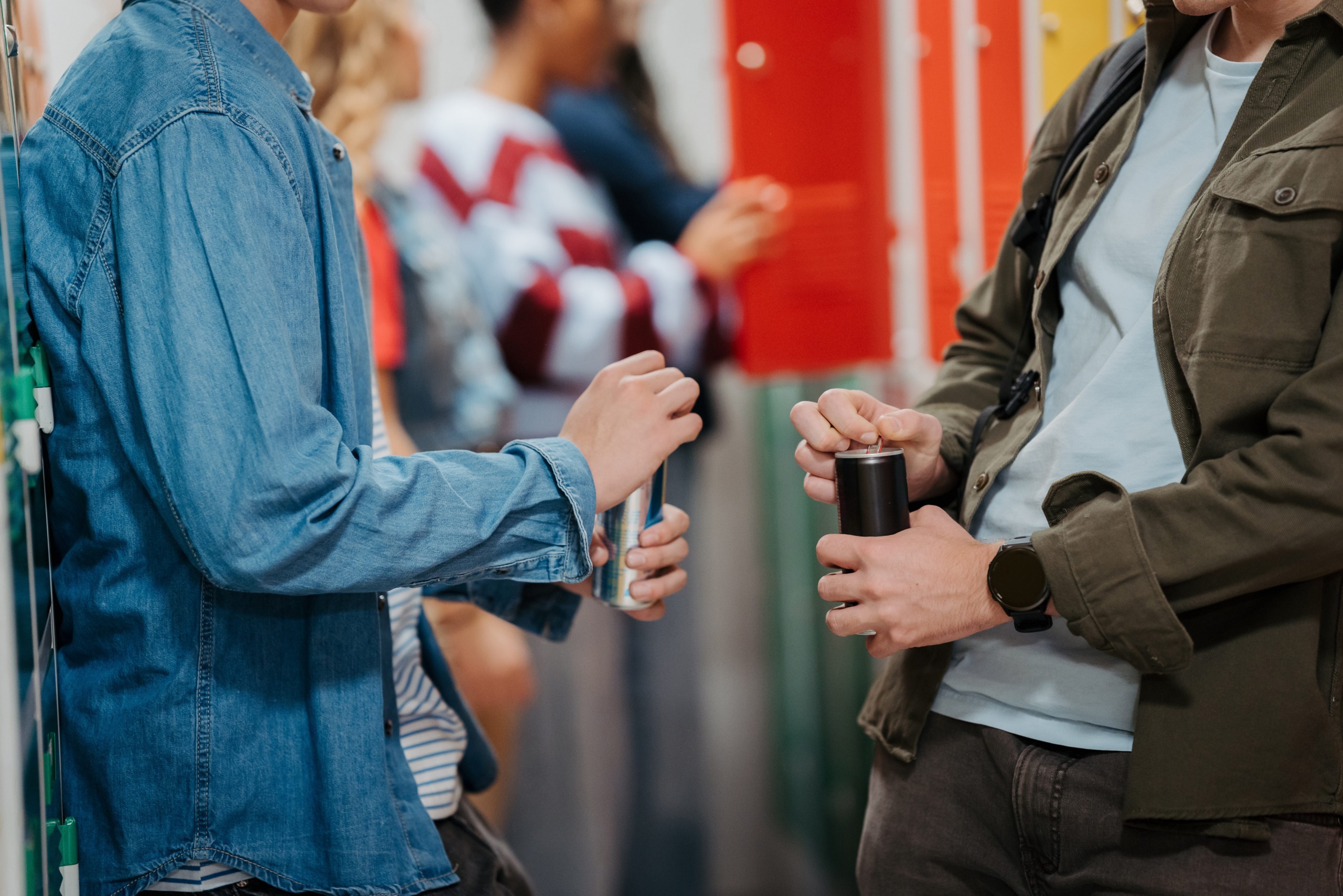 Two teenage students standing in a school hallway, both opening energy drink cans.